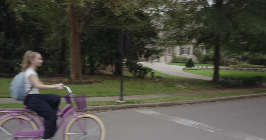 This is a shot of a suburban street in a leafy neighborhood on an overcast daya young man and a young girl ride bicycles toward the camera and then pass showing large trees and wellmaintained houses in the background