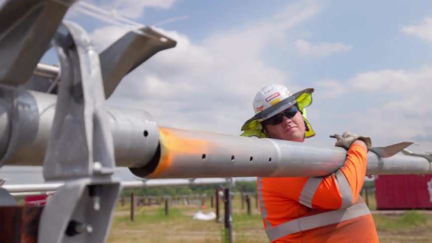 Worker in reflective orange shirt holds metal pipe at outdoor site with aligned structures
