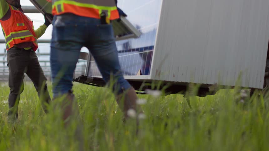 This is a lowangle closeup shot of a group of construction workers in safety vests installing large solar panels on a sunny daythe workers are seen lifting and maneuvering the panels beneath a partially installed row with the view looking up towar