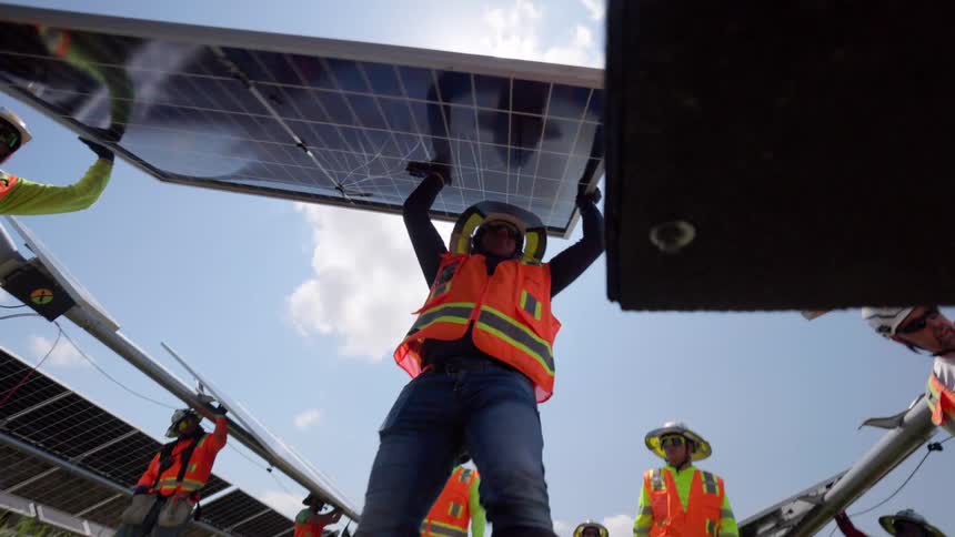 Multiple solar workers wearing orange safety vests hard hats and widebrimmed sun hats are installing a solar panel the main subject holds a panel overhead viewed from a low angle against the bright sun and a blue sky