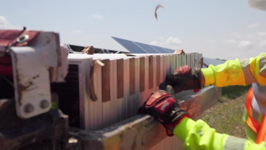 Worker in orange vest and hard hat handles metal structures beside solar panels under clear sky