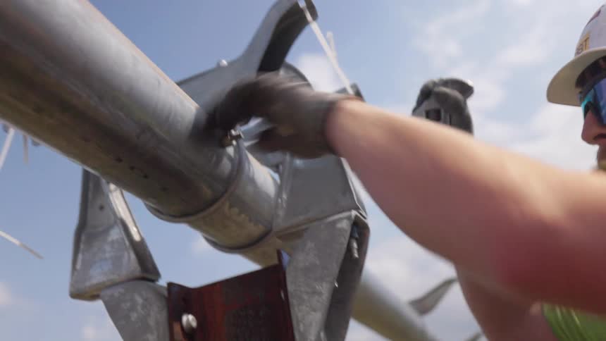 A worker wearing a hardhat and an orange vest uses a tool to secure a pole at a site midday