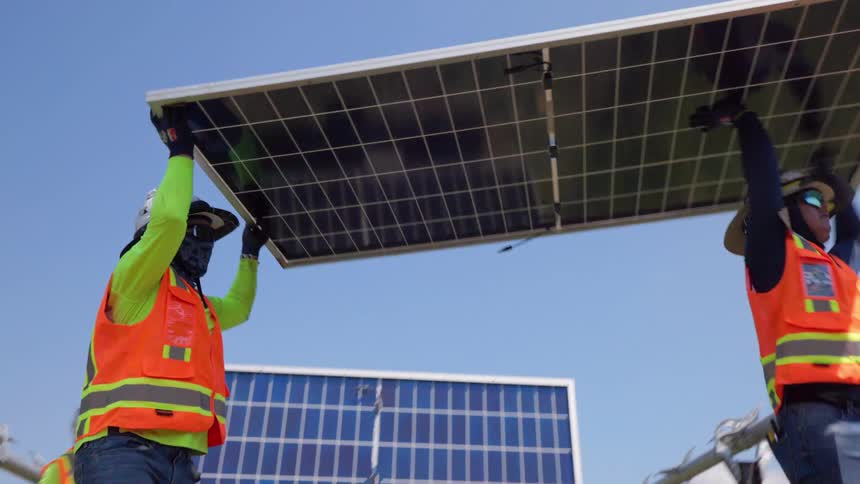 Multiple workers wearing safety vests and hats are shown collaboratively installing large solar panels onto a structure outdoors the bright sun and a clearpartially cloudy sky are visible as the panels are lifted into place