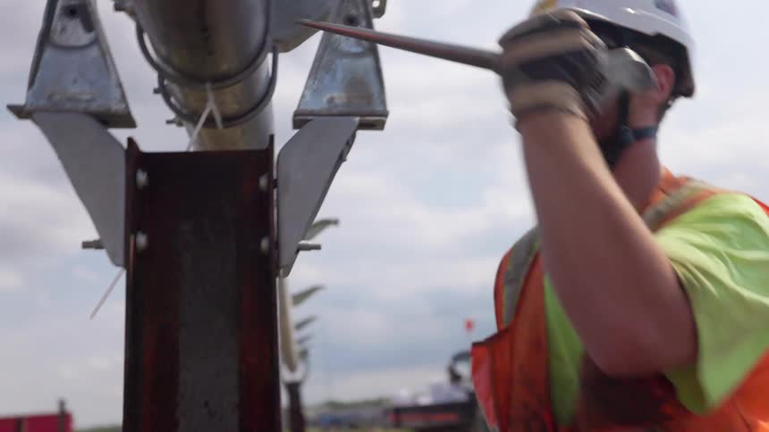 A construction worker wearing a white hard hat safety vest and sunglasses is shown tightening a bracket or clamp onto a large metal pipe or pole the work is being done outdoors under a partially cloudy sky