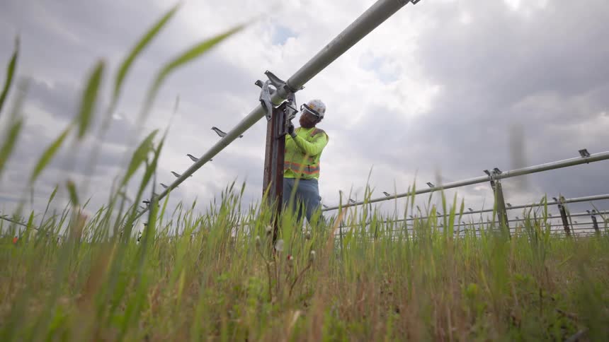 Worker in helmet adjusts metal irrigation structure in grassy field under cloudy sky