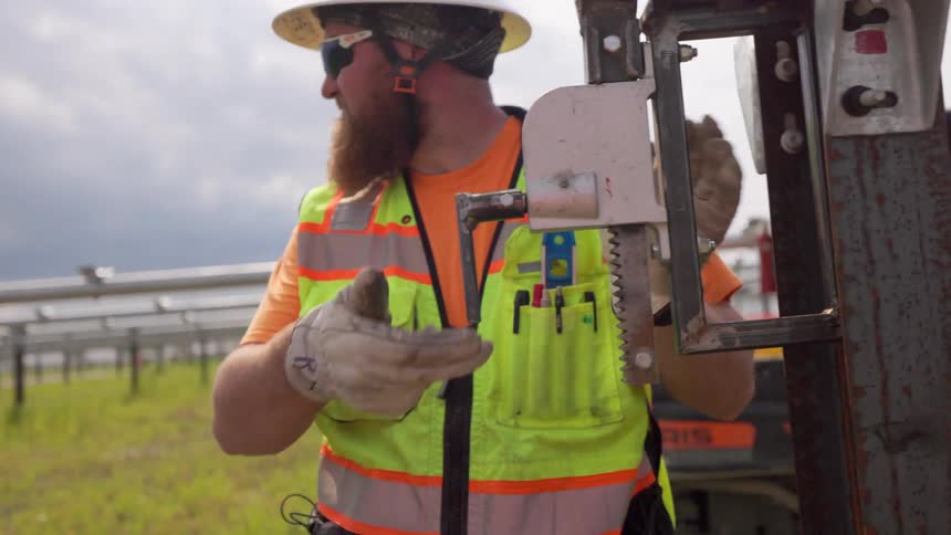 Worker in reflective vest adjusts serrated metal tool at construction site with vehicles in background