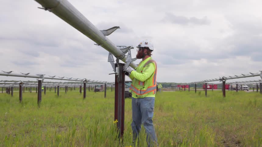 Construction worker wearing a helmet and sunglasses working on a metal structure outdoors surrounded by other workers and vehicles during day time