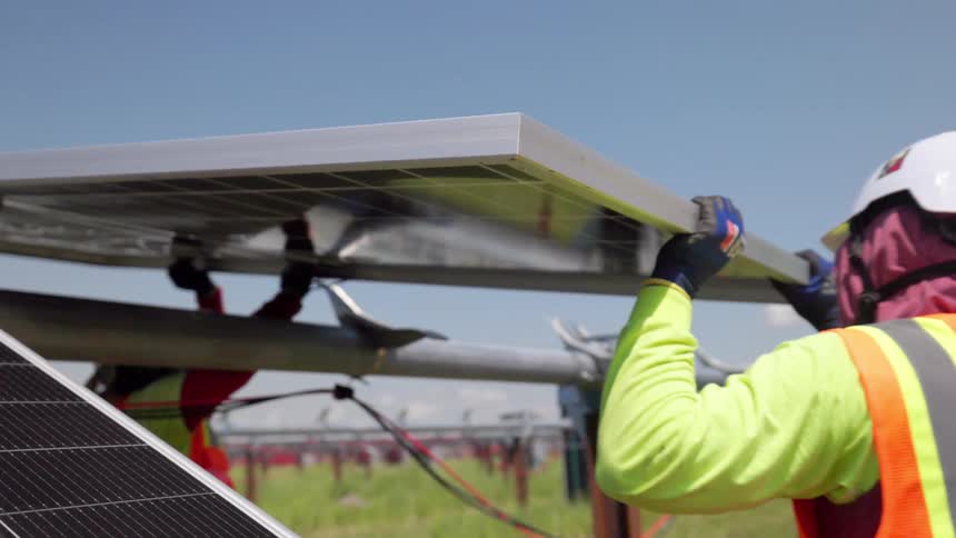 Two workers in orange vests adjust solar panel on metal structure under blue sky in open field