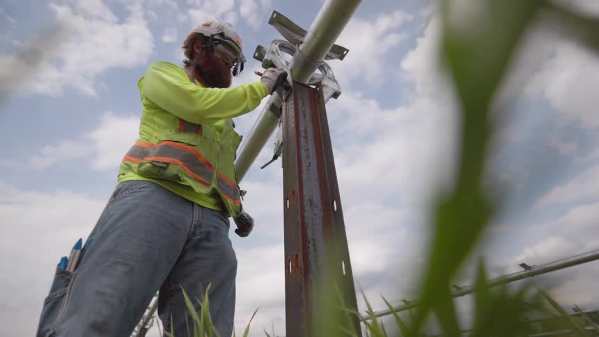 A worker in a hi vis vest uses a wrench to tighten a metal clamp on a vertical utility pole filmed from a low angle surrounded by tall green grass