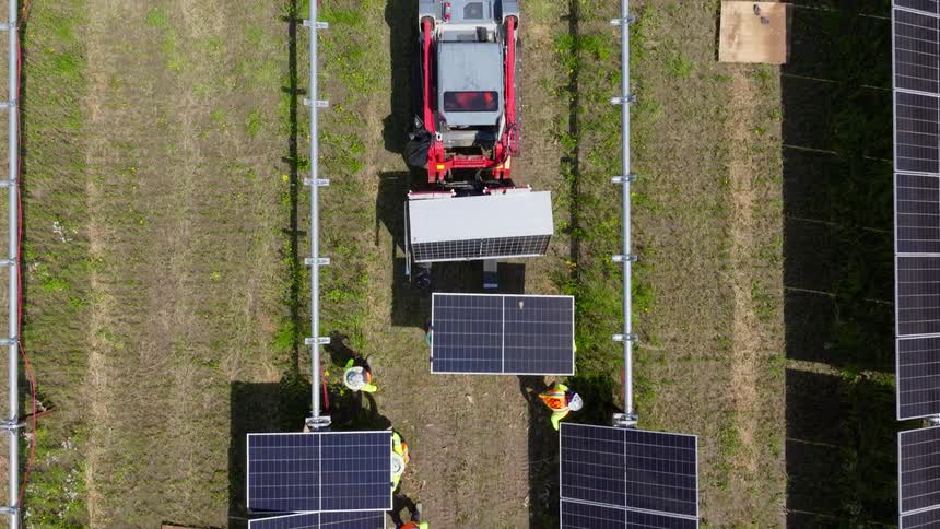 Red utility vehicle with solar panel in front is surrounded by workers installing structures in grassy field