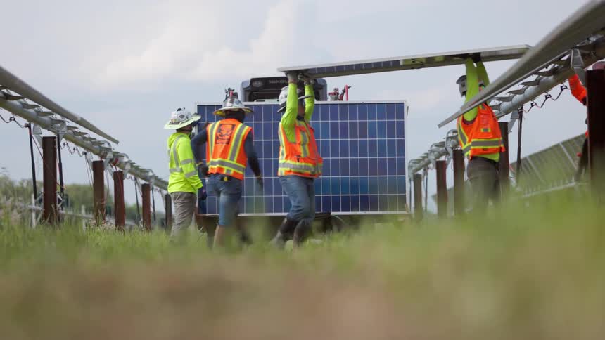 Workers with helmets and vests position solar panel between metal structures in grassy field under partly cloudy sky