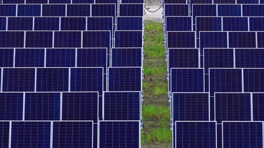 A slow aerial shot pans across endless rows of deep blue solar panels creating a vast geometric pattern over a green energy farm