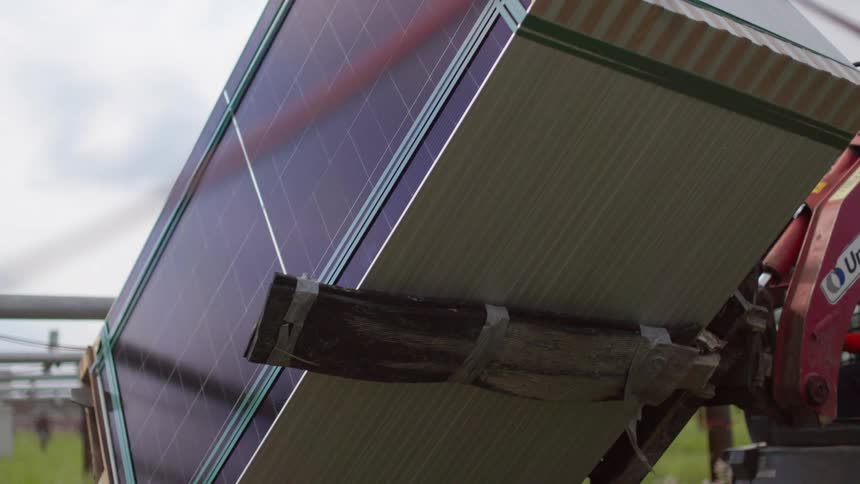 A machine tilts a large stack of solar panels held by a mechanical arm