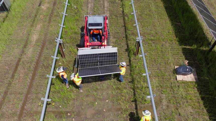 An aerial view shows workers unloading solar panels from a tracked carrier to install them on the metal racking system of a solar farm