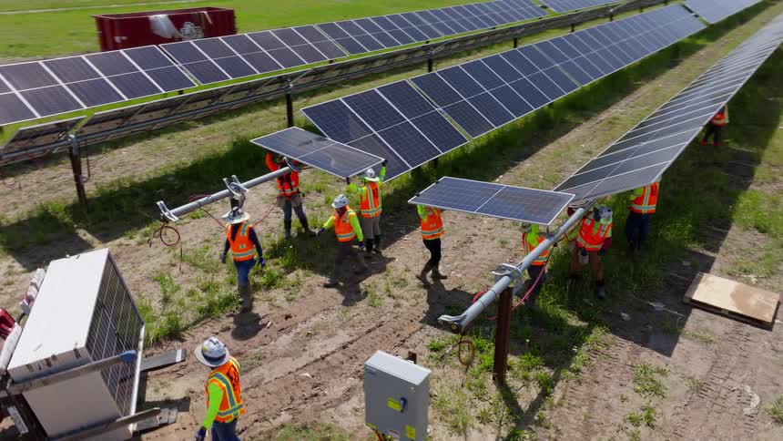Workers in vests and helmets install solar panels in grassy field with vehicles in background
