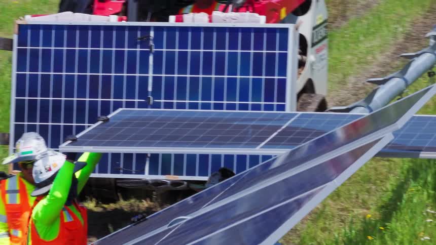 A group of workers wearing helmets and orange vests set up a series of solar panels in a solar field on a sunny day