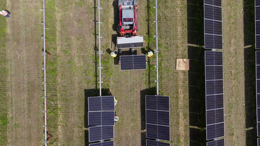 Workers in vests and helmets install solar panels in grassy field with vehicles in background