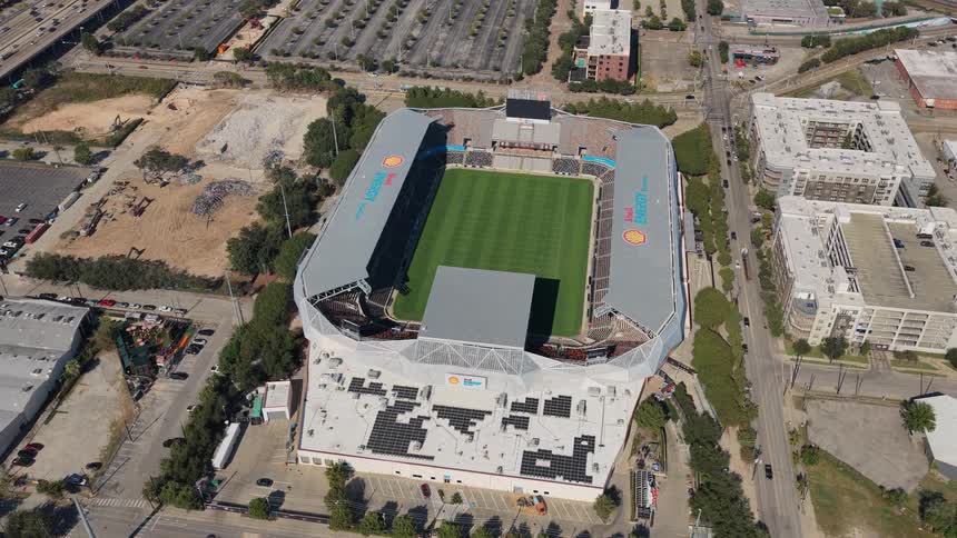 A slowpulling aerial shot reveals a modern sports stadium nestled within a bustling city surrounded by parking lots and urban development