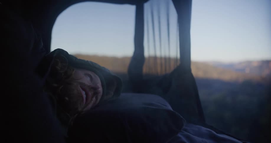 A man stirs from sleep inside a tent as soft morning light reveals a beautiful treecovered mountain range through the open flap