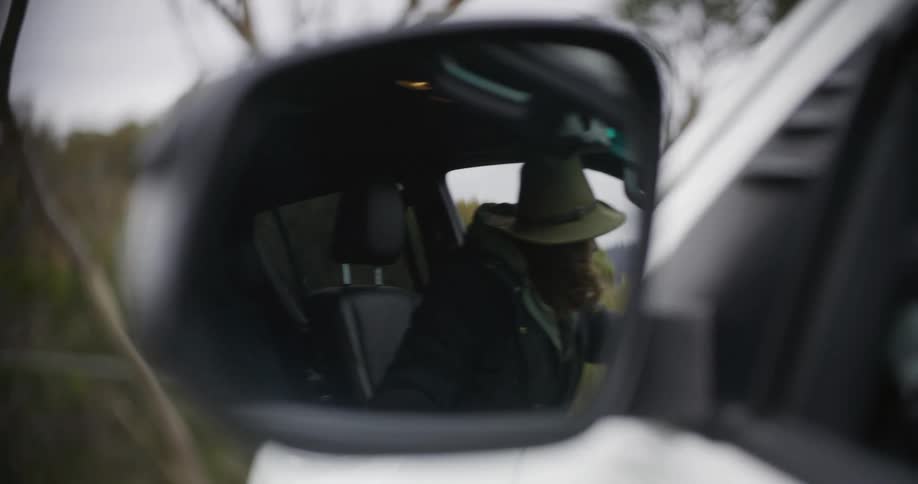 A young man with long hair and a beard walks up to his car gets in and shuts the door seen through the side mirror at midday