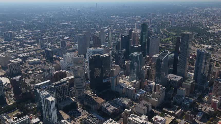 An expansive aerial view reveals a sprawling modern metropolis with its dense core of skyscrapers stretching towards a hazy horizon under the bright sun