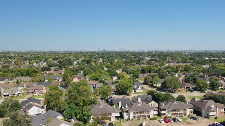 An aerial wide shot of a sprawling residential neighborhood characterized by dense tree cover and houses under a bright blue sky the distant skyline of a major city is visible on the horizon suggesting the suburb is located on the outskirts of an urba