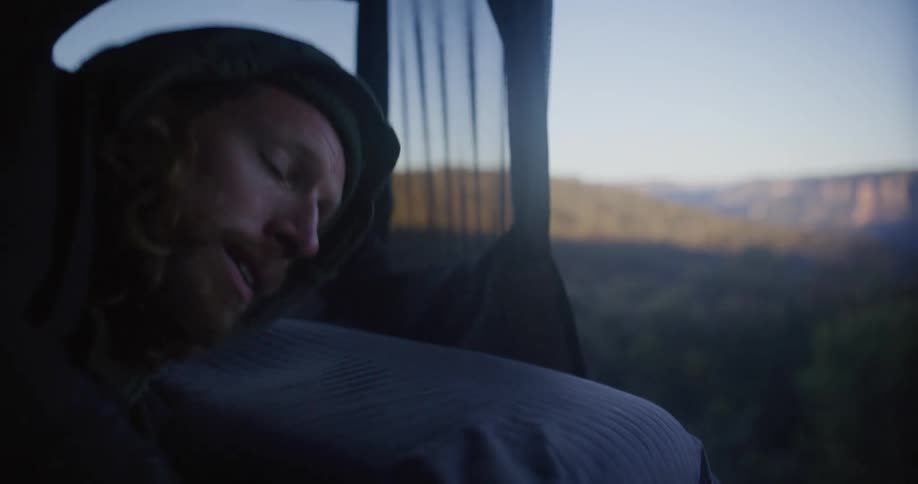 A man wakes up inside a tent revealing a scenic view of a forested canyon landscape at sunrise