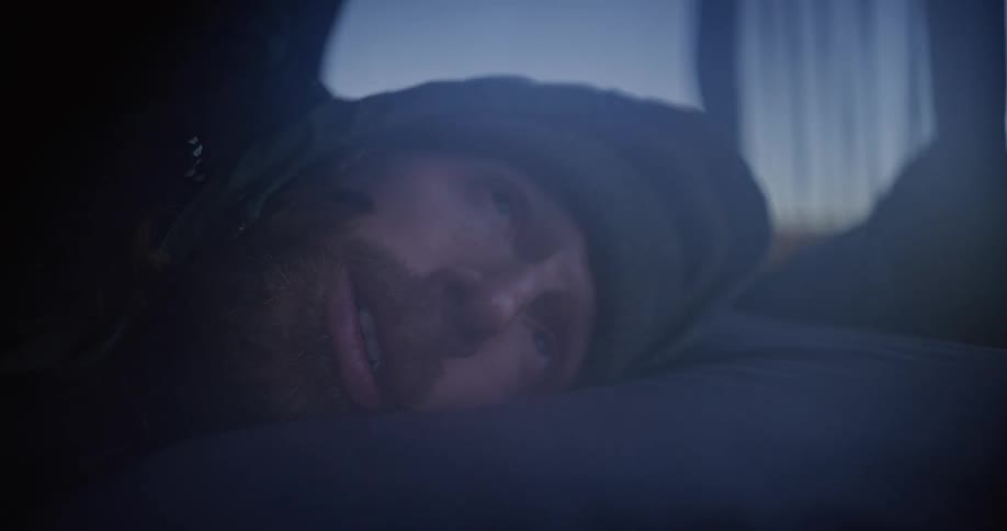 The face of a man bundled in a sleeping bag and a beanie is shown in a close view while resting inside a dark tent at dawn