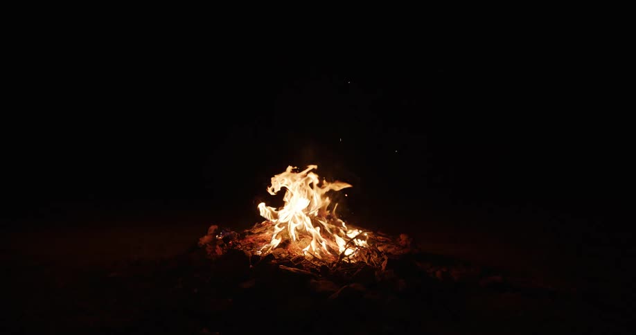 Campfire burning on the ground with logs igniting and sparks rising into the air