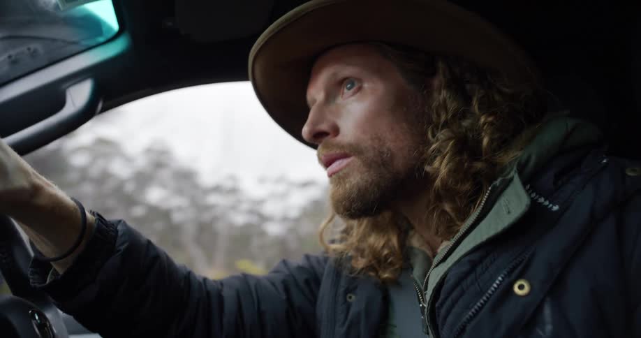 A young man with long hair facial hair and a hat drives a truck on a countryside freeway at midday