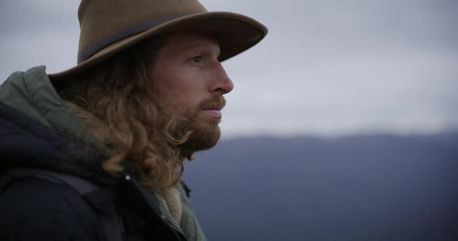 Person observes mountainous landscape under cloudy sky with backpack and widebrimmed hat