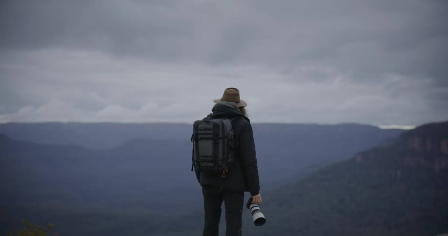 A person wearing a hat black jacket and backpack stands on a lookout point gazing at a vast misty landscape of rolling hills and valleys under a gray sky they are holding a large camera lens in their hand indicating they are likely a photographer or