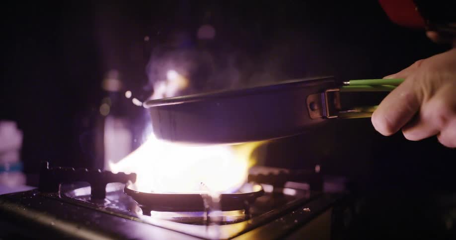 High flame rising beneath a frying pan held by a hand over a portable stove