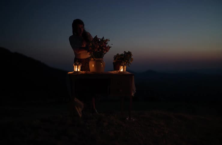 A woman is dimly lit by two flickering lanterns as she stands over a small table on a grassy hilltop at twilight she is adjusting a large vase filled with flowers silhouetted against the dark blue hues of the sky and distant hills