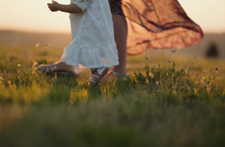 Child in a white dress walking on the grass beside an adult during sunset