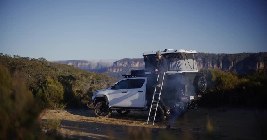 A man sits in the doorway of a rooftop tent mounted on a white truck overlooking a scenic canyon landscape