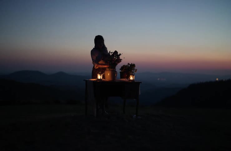 A woman is seen outdoors on a hillside at twilight carefully arranging flowers in a large ceramic pot on a small table lit by two lanterns in a contrasting scene the same woman walks and kneels beside a sunny rocky stream in a forest dipping her hand