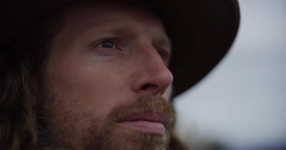 Closeup of a male face with light beard and hat framed against a cloudy sky
