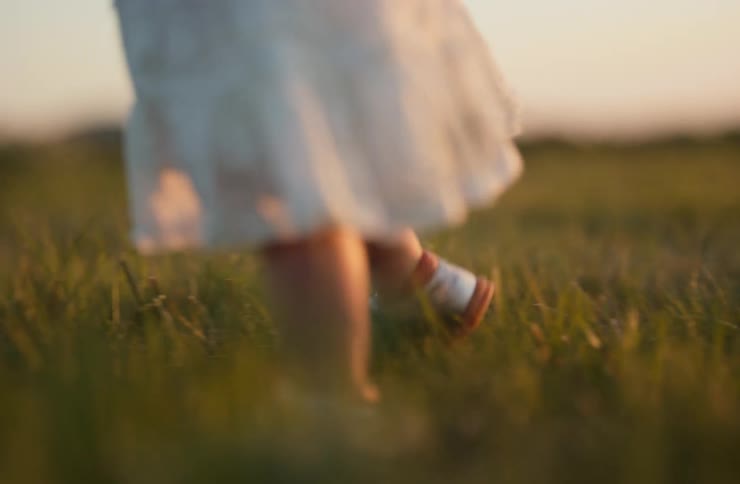 A toddler wearing a dress and sandals walks across a grass field at sunset