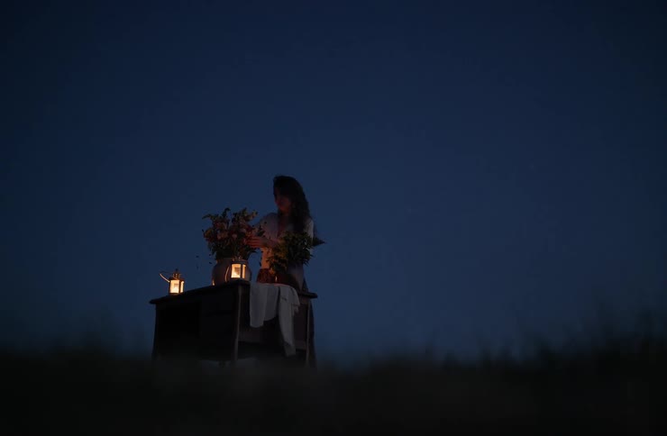 Woman stands behind table decorated with flowers and lit lanterns during nighttime