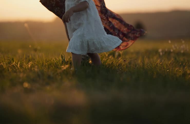 Child in a white dress walking on the grass beside an adult during sunset