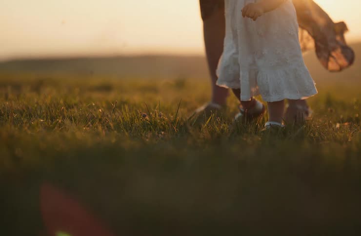 Child in a white dress walking on the grass beside an adult at sunset