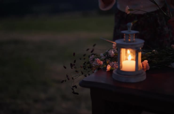 A lit lantern glows on a table while a person arranges flowers in the background