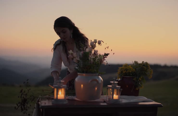 A woman stands behind a table on a hillside arranging a large bouquet of flowers in a pot silhouetted against a colorful sunset two lit lanterns flank the centerpiece casting a warm glow over the rustic setting