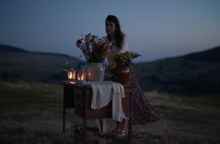 A woman arranges flowers on a wooden desk illuminated by lanterns in a grassy meadow at dusk
