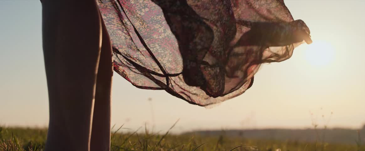 A close view of a sheer floral printed womans skirt gently blowing in the wind brightly backlit by the setting sun in a grassy field