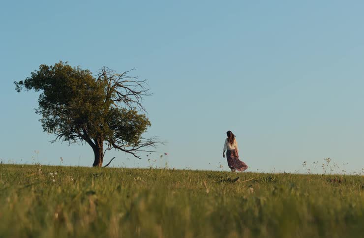 A woman in a skirt is walking in a field with lots of grass and a small tree