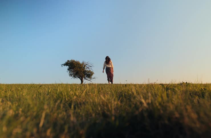 Woman wearing a long skirt walks across a rural field toward a tree