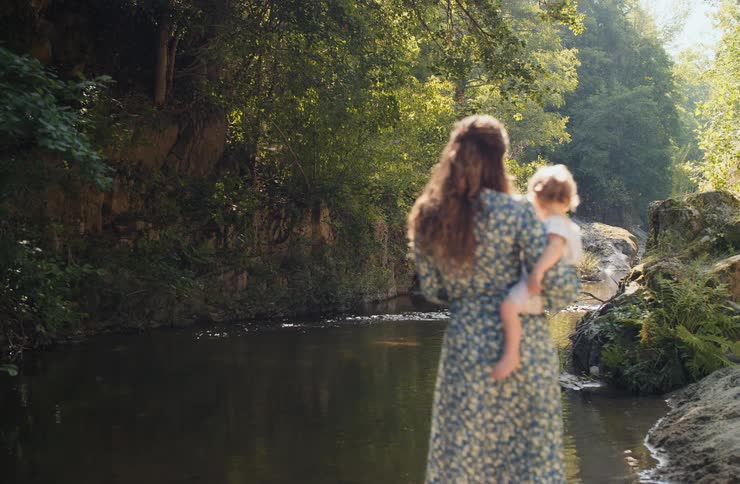 Woman holds child near stream in wooded area under soft natural light