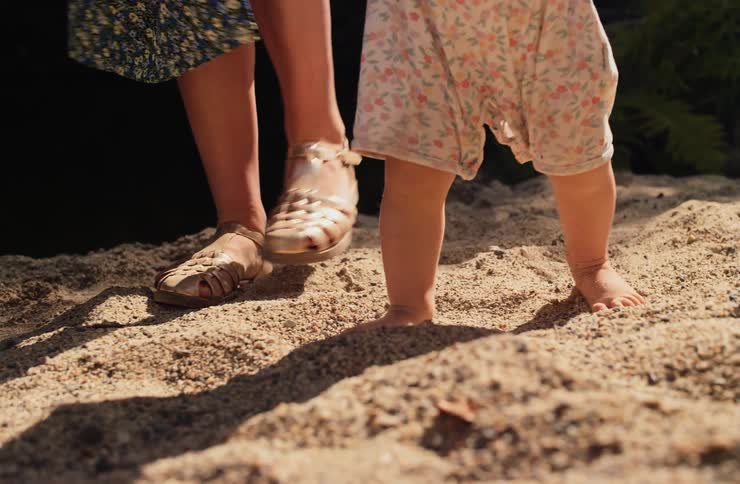 In the warm sunlight a toddler takes wobbly steps barefoot through the sand as their mother stands patiently beside them